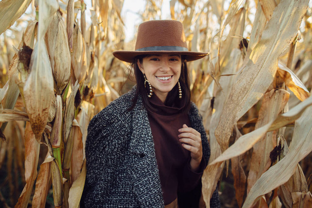 Portret szczęśliwej stylowej kobiety w brązowym kapeluszu i płaszczu vintage spaceru w jesiennym polu kukurydzy w świetle zachodu słońca. Modna młoda hipsterka w stroju retro uśmiechnięta w jesiennej kukurydzy - Zdjęcie, obraz