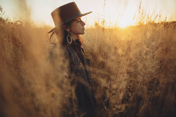 Красива стильна жінка в капелюсі і вінтажне пальто, що стоїть в траві в теплому світлі заходу сонця в осінньому полі. Спокійний атмосферний момент. Модна молода жінка насолоджується вечором у сільській місцевості
 - Фото, зображення
