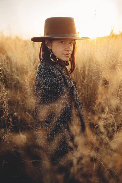 Mulher bonita e elegante em chapéu e casaco vintage em pé na grama em luz quente do pôr do sol no campo de outono. Momento atmosférico tranquilo. Elegante jovem do sexo feminino desfrutando de noite no campo - Foto, Imagem