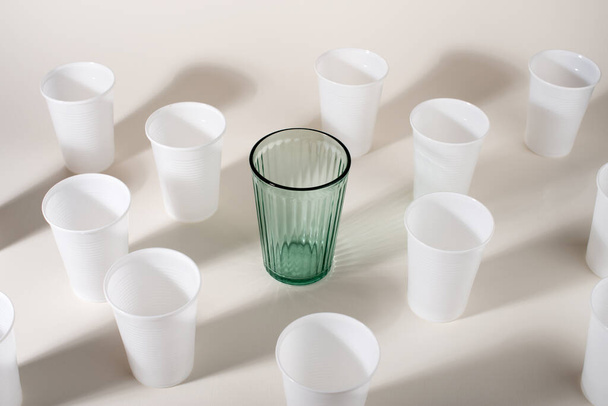 多くのプラスチックカップの間でガラス。ガラスかプラスチックだ。再利用可能と使い捨て。廃棄物ゼロのコンセプト。プラスチック汚染を止める. - 写真・画像