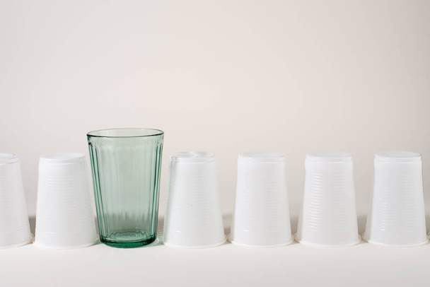 生のガラスと多くのプラスチックカップ。ガラスかプラスチックだ。再利用可能と使い捨て。廃棄物ゼロのコンセプト。プラスチック汚染を止める. - 写真・画像