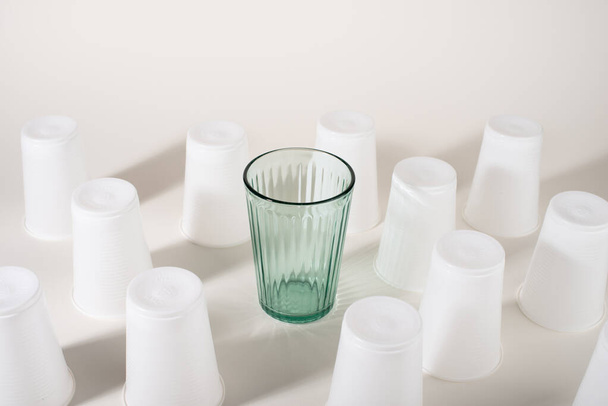 多くのプラスチックカップの間でガラス。ガラスかプラスチックだ。再利用可能と使い捨て。廃棄物ゼロのコンセプト。プラスチック汚染を止める. - 写真・画像