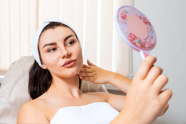 Porträt einer kaukasischen Frau, die sich selbst im Spiegel betrachtet, in einem Schönheitssalon. Konzept der Kosmetologie und plastischen Chirurgie. - Foto, Bild