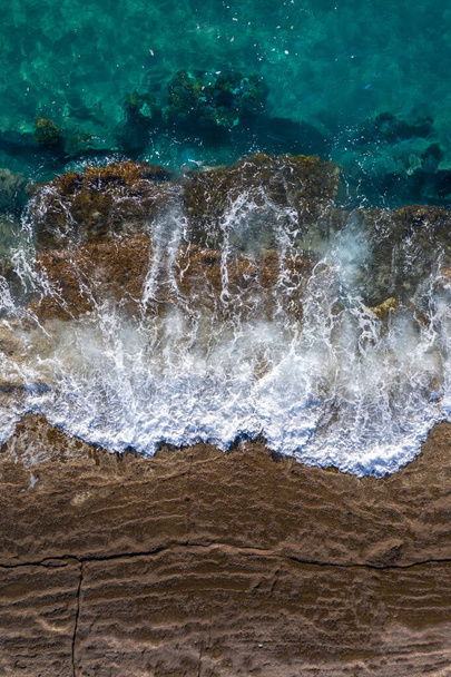 Waves breaking on brown cliff rocks with rising tide, drone perspective directly above - Photo, Image