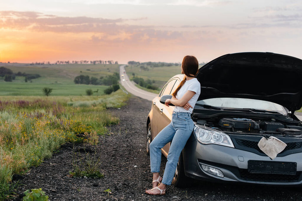 A frustrated young girl stands near a broken-down car in the middle of the highway during sunset. Breakdown and repair of the car. Waiting for help. - Foto, Bild