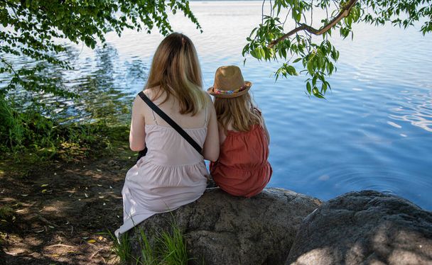 deux filles sont assises sur un rocher sous un arbre sur la rive d'un lac par une journée d'été ensoleillée. Sans visage - Photo, image