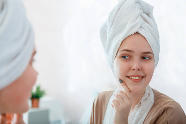 Self Care as part of morning bathroom routine. Portrait of smiling young woman with perfect skin in mirror reflection after taking shower before makeup with towel on head. - Foto, afbeelding