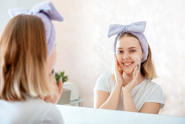 Teen girl washing face portrait reflection in mirror in morning bathroom. Young blonde woman applies facial Cleaner for skin wash. Self Care morning bathroom routine - Zdjęcie, obraz