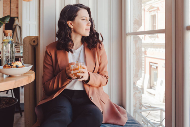 Улыбающаяся женщина сидит в кафе и пьет чай, глядя в окно. - Фото, изображение