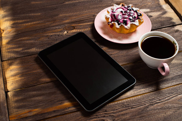 Διάταξη διαλείμματος καφέ Flatley. Ψηφιακό tablet gadget, μαύρο καφέ σε ένα φλιτζάνι και ένα καλάθι κέικ με μούρα σε ένα τραπέζι από ένα παλιό σκούρο ξύλο. - Φωτογραφία, εικόνα