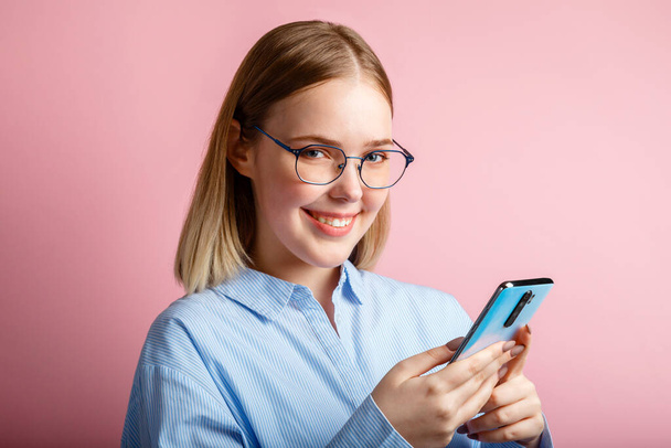 Emocjonalny portret szczęśliwego pracownika biura menedżera uśmiechu w okularach używać smartfona. Kobieta w niebieskiej koszuli pisze wiadomość przez telefon odizolowany na różowym tle. - Zdjęcie, obraz