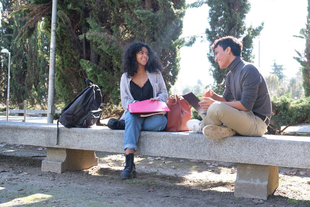 Двоє латинських студентів посміхаються, навчаючись разом, сидячи на лавці на відкритому повітрі. Університетське життя в кампусі
. - Фото, зображення