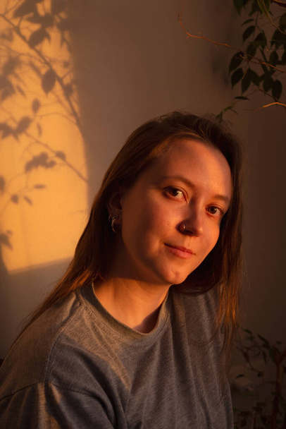 Young woman looking at camera in warm rays of sunset. Warm calm portrait of girl indoor. light and shadow on the face - Foto, afbeelding