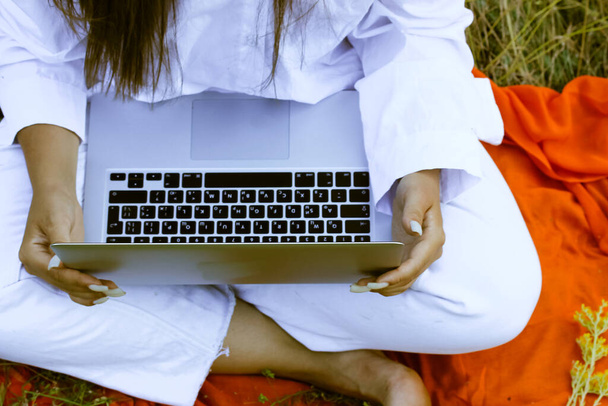 An unrecognizable girl in a white suit is typing on a laptop keyboard sitting on an orange plaid in nature. Yellow flowers lie on the computer. Online business, communication, study, work concept.  - Photo, Image