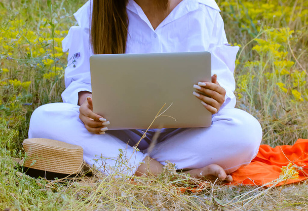 Une fille méconnaissable en costume blanc tape sur un clavier d'ordinateur portable assis sur un plaid orange dans la nature. Fleurs jaunes gisent sur l'ordinateur. Affaires en ligne, communication, étude, concept de travail.  - Photo, image