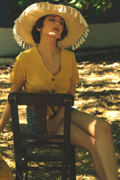 Country woman sits on a wooden chair with a straw hat yellow shirt and jean shorts. - Fotó, kép