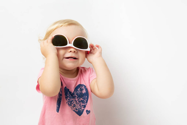 Portrait of cheerful curly little baby girl in a pink t-shirt wearing sunglasses and looking away on white background with copy space - Photo, Image