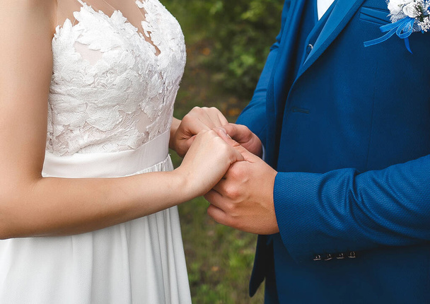 Ο γαμπρός με το μπλε κοστούμι κρατάει τα χέρια της νύφης με ένα λευκό φόρεμα. Ευαισθησία και στοργή στο γάμο. - Φωτογραφία, εικόνα