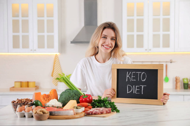 Donna con lavagna con frase Keto Diet e diversi prodotti a tavola bianca in cucina - Foto, immagini