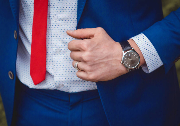 The hand of a man with a hand watch holds on to a blue suit against the background of a white shirt and red tie, a business men's style of clothing decor. - Fotografie, Obrázek