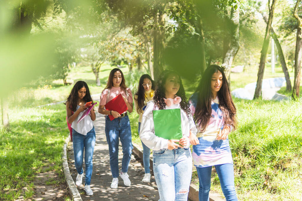 Foto di due ragazze adolescenti latine che camminano tra gli alberi per andare a lezione in un campus con tre ragazze fuori fuoco dietro - Foto, immagini