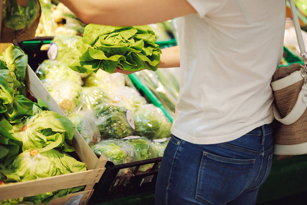 Nuori nainen valkoisessa t-paidassa ja laukku olkapäällään valitsee salaatin vehreyden osastolta. Luonnonmukaista ruokaa ja vitamiineja. Oikea ravitsemus. Ruokavalio. Maku ja nautinto. Kesäruoka. - Valokuva, kuva