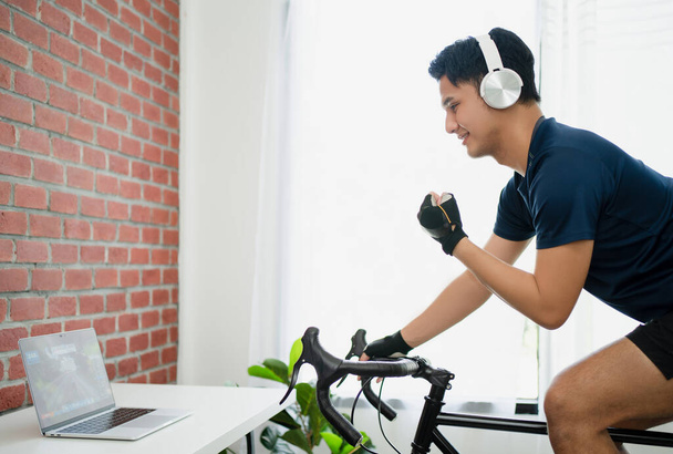 Conceptos de salud y estilo de vida. Hombre asiático cruza la línea de meta en una carrera jugando juegos de bicicleta en línea en el ordenador portátil. Él ciclismo en el entrenador en la casa. - Foto, Imagen