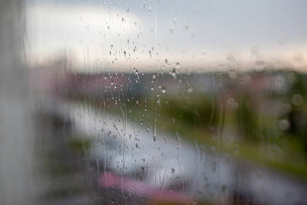 Regenachtige dag door het raam op bewolkte grijze lucht en stadsgebouwen achtergrond. Concept. Avond stadsgezicht achter het raam met druppelende druppels water. - Foto, afbeelding