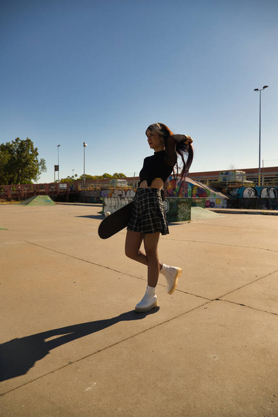 Young and pretty girl with punk style with a skateboard under her arm holding a ponytail in a skateboard park. Vertical position. - Zdjęcie, obraz