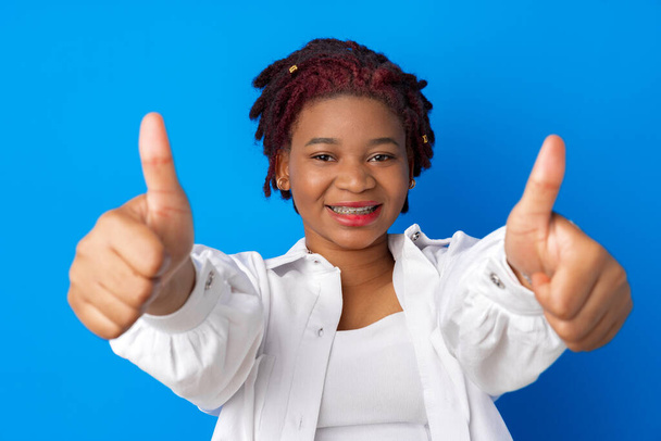 Улыбающаяся молодая афро-женщина показывает большой палец вверх знак на синем фоне - Фото, изображение