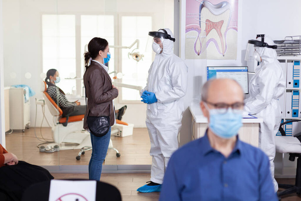 Οδοντίατρος ντυμένος με κοστούμι ppe συζητά με τον ασθενή - Φωτογραφία, εικόνα