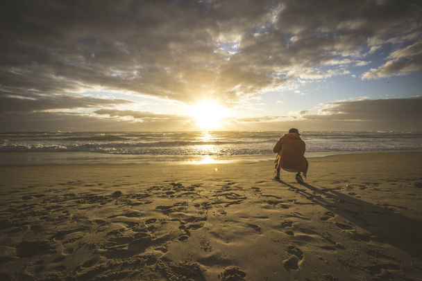 A squatted man on the sandy beach taking a shot of a sunset - Fotoğraf, Görsel