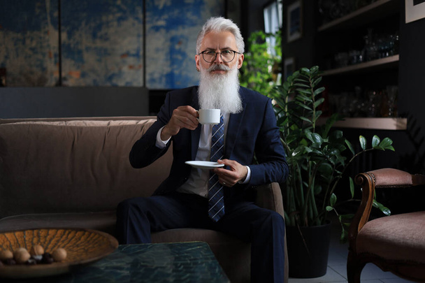 Handsome mature man holding coffee cup sitting on the sofa in office. Coffee break - Фото, изображение