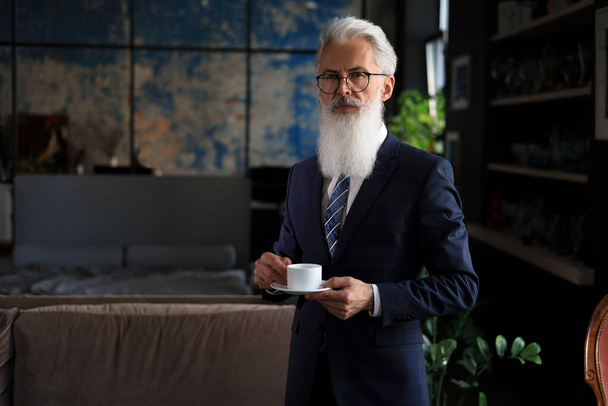 Start of a new working day with a cup of coffee. Handsome mature man holding coffee cup standing in his modern office - Photo, Image