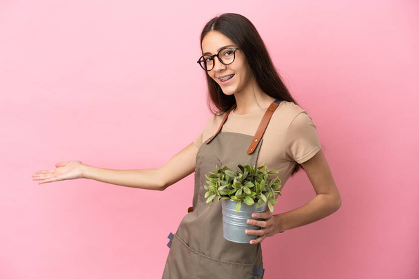 Joven jardinero sosteniendo una planta aislada sobre fondo rosa extendiendo las manos a un lado para invitar a venir - Foto, imagen