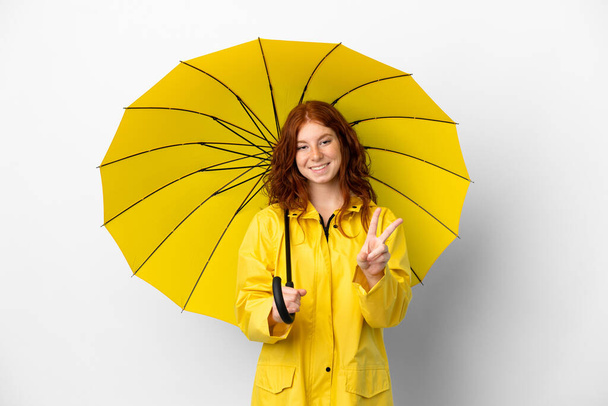 Adolescente ruiva menina capa impermeável e guarda-chuva isolado no fundo branco sorrindo e mostrando sinal de vitória - Foto, Imagem