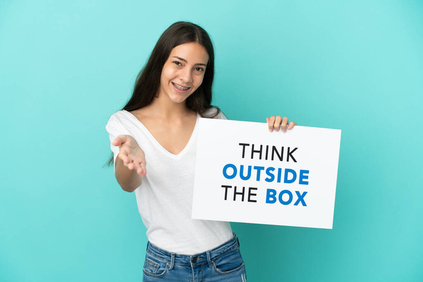 Молодая француженка, изолированная на синем фоне, держит плакат с текстом "Думай вне коробки" и заключает сделку. - Фото, изображение
