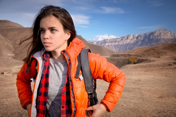 Portrait of a young Caucasian attractive woman standing in a backpack on the background of a rocky mountain peak looks to the side while adjusting her jacket - Photo, Image