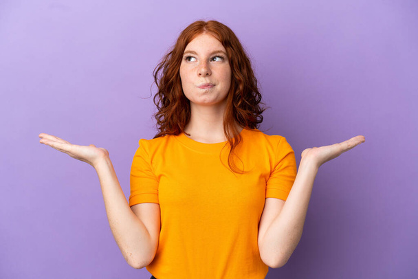 Teenager redhead girl over isolated purple background having doubts while raising hands - Foto, imagen