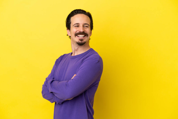 Young caucasian man isolated on yellow background with arms crossed and happy - Fotografie, Obrázek