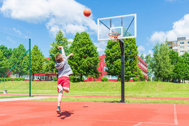 Attrayant jeune garçon tir balle au cerceau à playground.cute jeune garçon joue au basket-ball sur l'aire de jeux dans la journée chaude d'été. - Photo, image