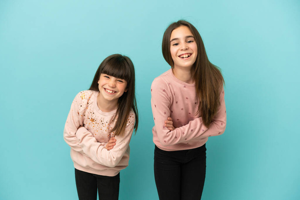 Kleine zusjes meisjes geïsoleerd op blauwe achtergrond houden de armen gekruist terwijl ze glimlachen - Foto, afbeelding