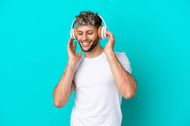 Genç yakışıklı beyaz adam mavi arka planda izole edilmiş müzik dinliyor ve şarkı söylüyor. - Fotoğraf, Görsel