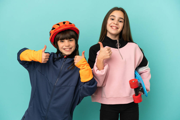 Siostry ćwiczące jazdę na rowerze i jazdę na łyżwach odizolowane na niebieskim tle dające kciuki w górę gestem obiema rękami i uśmiechnięte - Zdjęcie, obraz