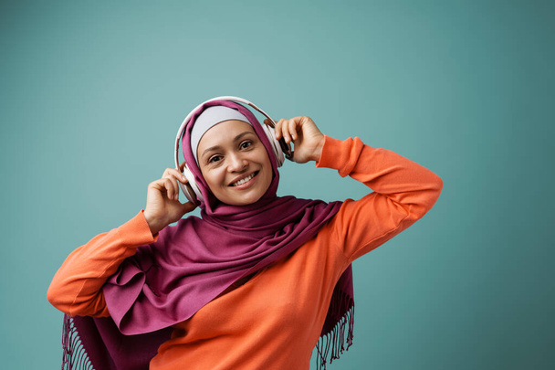 Middle eastern woman in hijab smiling and using headphones isolated over blue background - Φωτογραφία, εικόνα