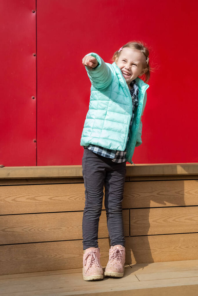 Adorable little caucasian girl sitting on a wooden deck next to a red wall looking at the camera - Foto, imagen
