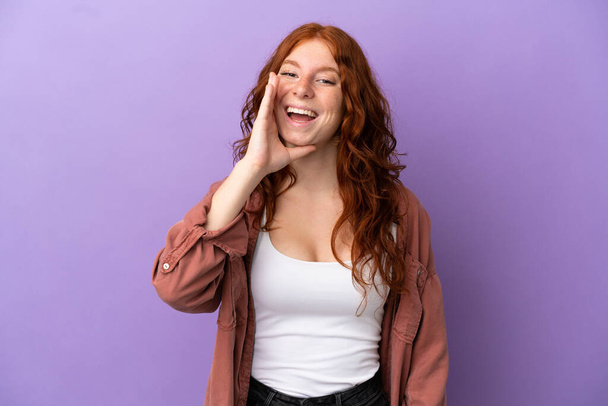 tiener roodharige meisje over geïsoleerde paarse achtergrond schreeuwen met mond wijd open - Foto, afbeelding