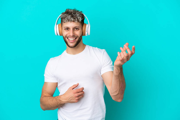 Молодой красивый белый мужчина изолирован на синем фоне слушает музыку и делает гитарный жест - Фото, изображение