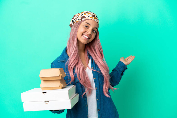 Young mixed race woman holding pizzas and burgers isolated on green background extending hands to the side for inviting to come - Foto, Imagem