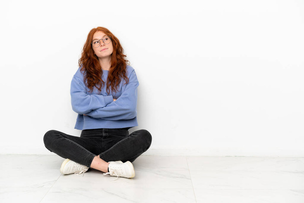 Подросток рыжая девушка сидит на полу изолированы на белом фоне делает сомнения жест, поднимая плечи - Фото, изображение